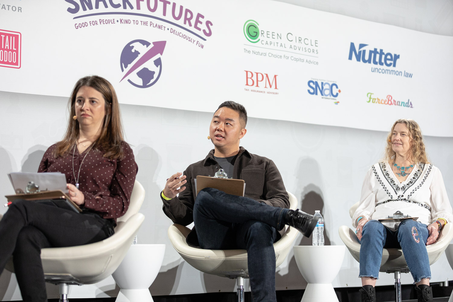 Woman in a burgundy polka-dot top, man in a brown jacket speaking and woman in a white embroidered blouse seated in white chairs in front of sponsor logos including SnackFutures, Green Circle Capital Advisors, Nutter, BPM and SNAC.