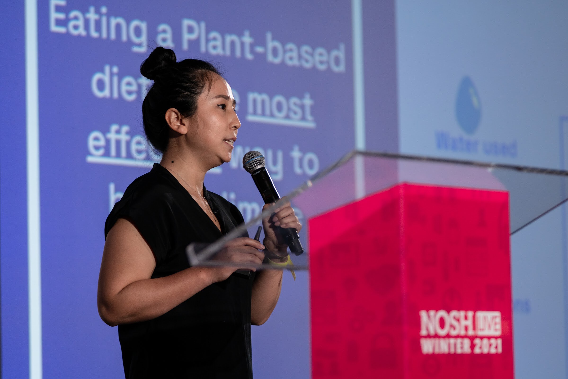 Woman with her hair in a bun holding a microphone at a clear podium with a pink base, in front of a blue slide reading Eating a Plant-based diet.