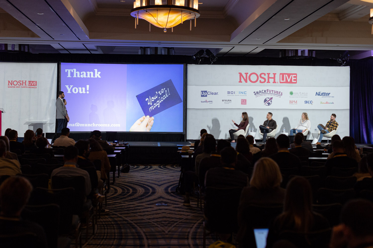 Wide view of the Nosh Live stage with a blue Thank You slide that includes a munchrooms email address, a presenter at the left and four judges seated at the right, with the audience in the foreground.