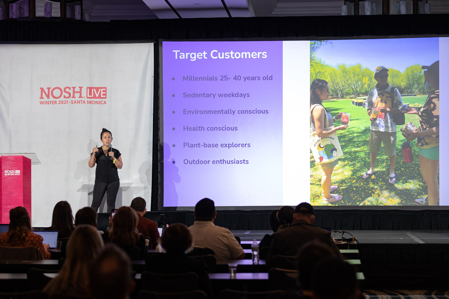 Woman in a black top on stage beside a pink podium, with a purple slide reading Target Customers and a photo of people holding Munchrooms products, and audience heads in the foreground.