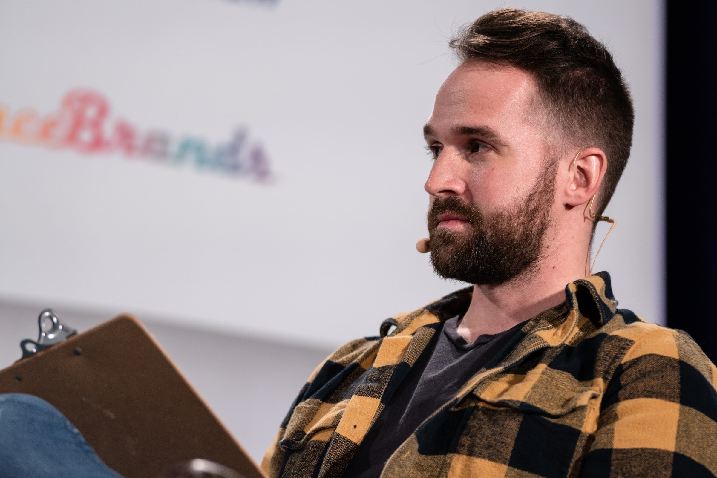 Close view of a bearded man in a yellow and black plaid shirt with a headset microphone looking to the side, a clipboard on his lap.