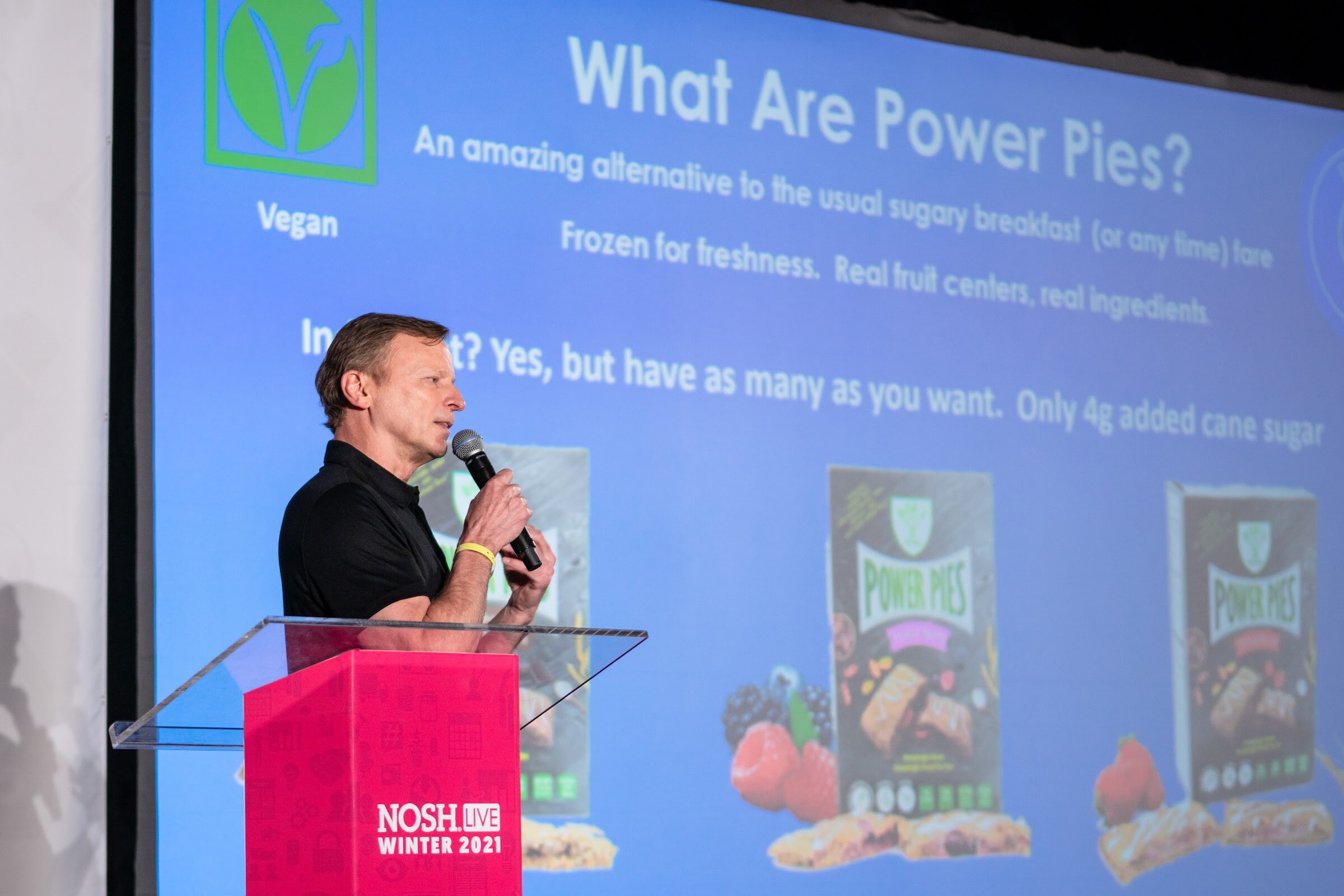 Man in a black polo with a microphone at a pink Nosh Live podium in front of a blue slide titled What Are Power Pies with pictures of Power Pies boxes.