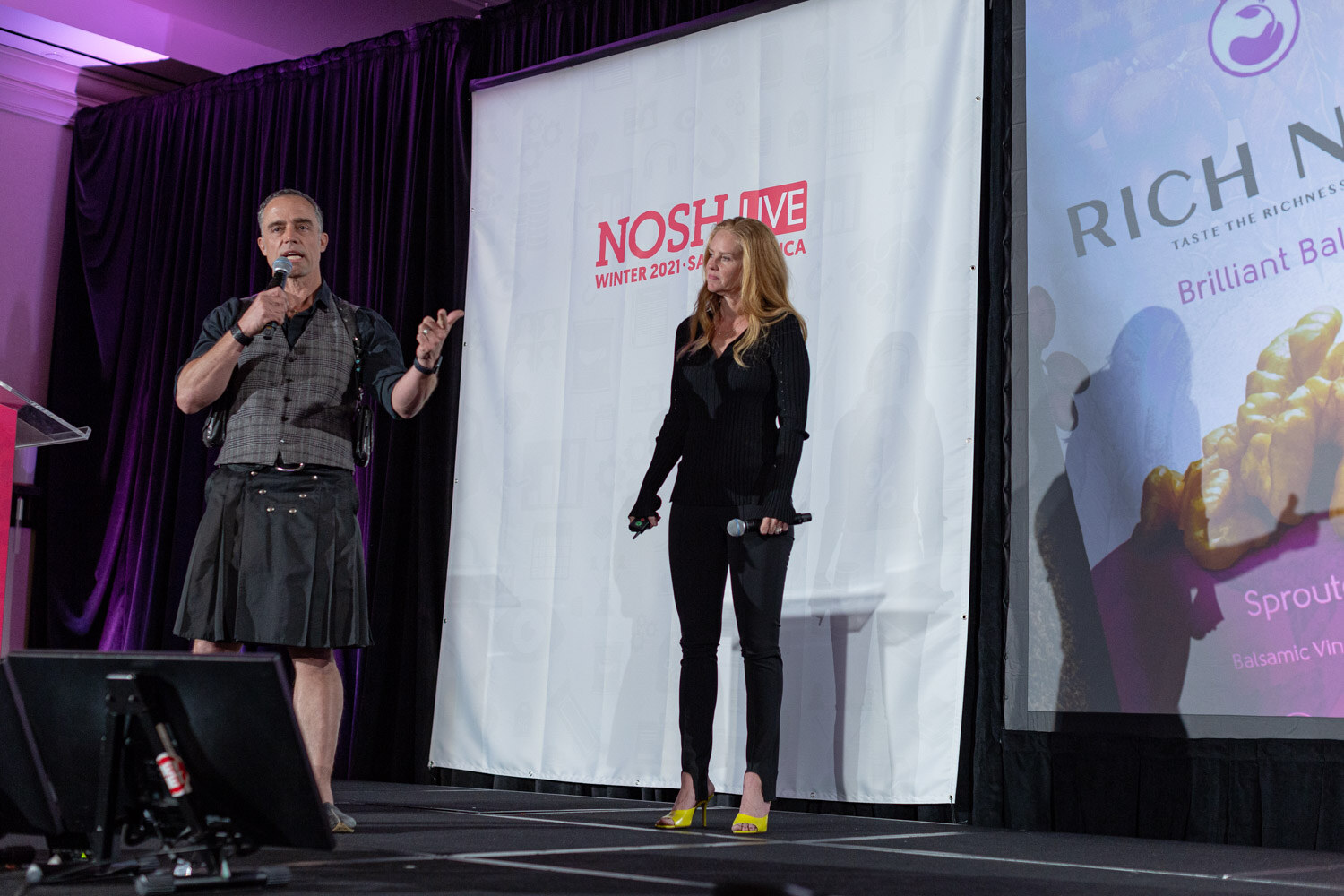 Man in a vest and kilt gesturing with a microphone and a tall woman in black standing on a stage in front of a Nosh Live banner and a Rich Nuts slide.