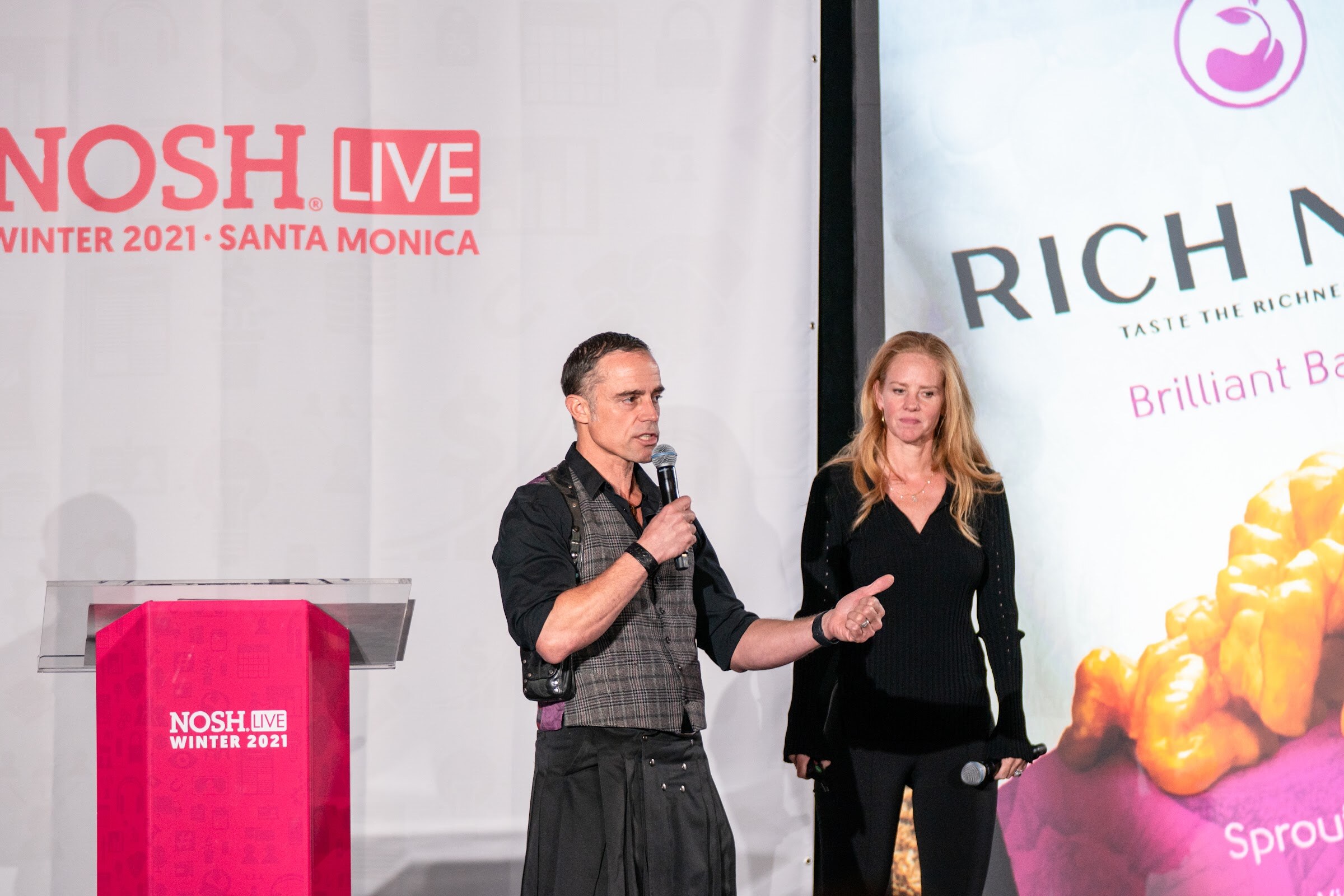 Man in a plaid vest and black kilt holding a microphone and a woman in black standing beside a podium, with a Rich Nuts slide at the right.