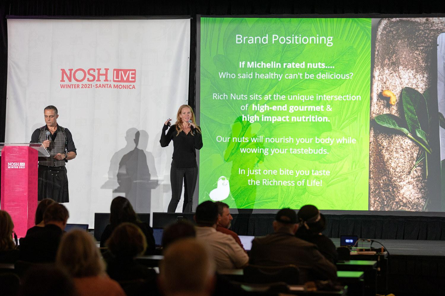 Man in a vest and kilt and a woman in black stand on stage beside a green slide titled Brand Positioning, with the audience in silhouette below.
