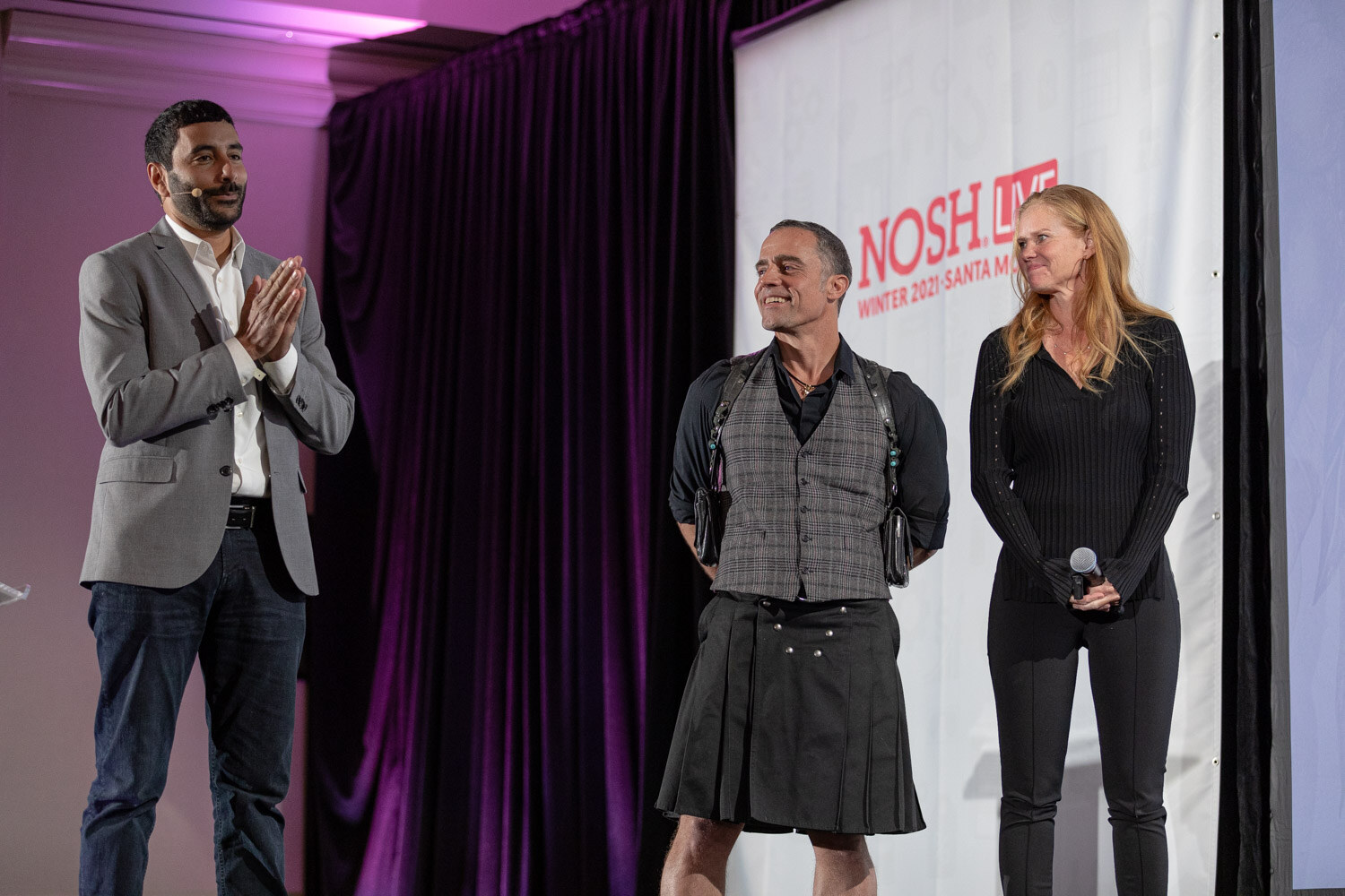 Man in a grey blazer clapping next to a man in a plaid vest and kilt and a woman in black, standing in front of a Nosh Live banner and purple curtain.