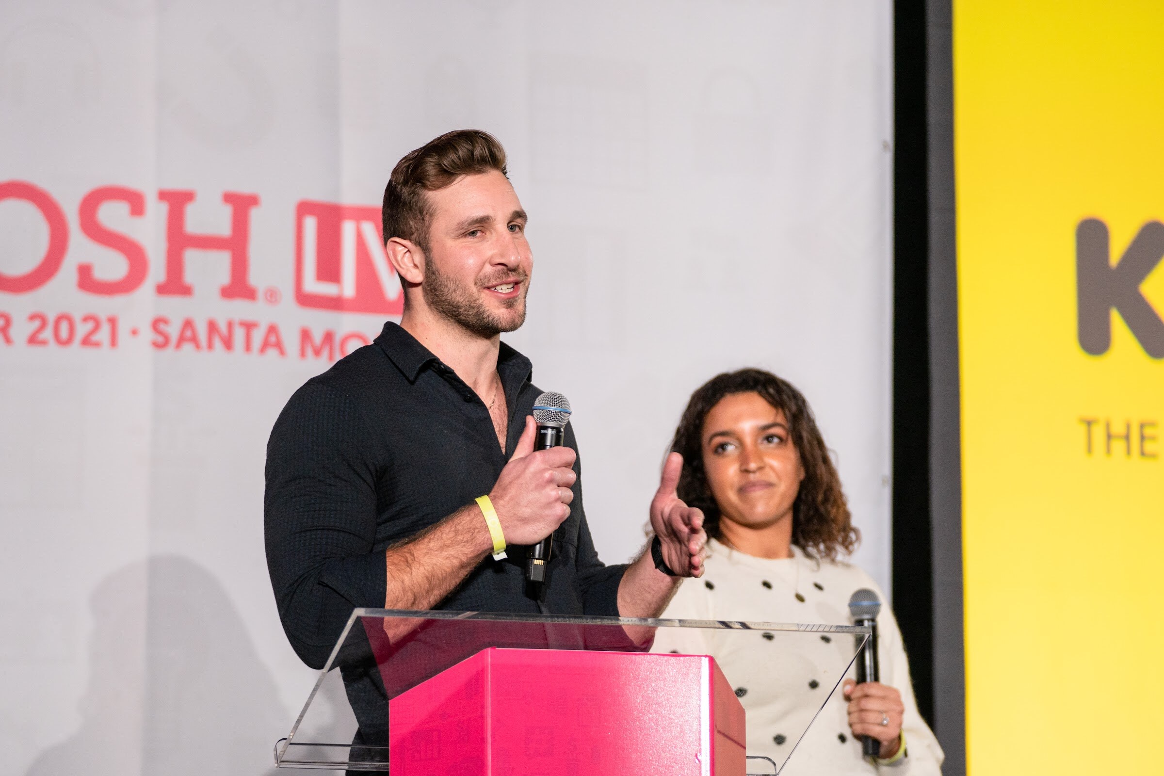 Man in a black button-up shirt holding a microphone at a podium and a smiling woman in a cream polka-dot sweater beside him, with a yellow Kokada slide behind them.