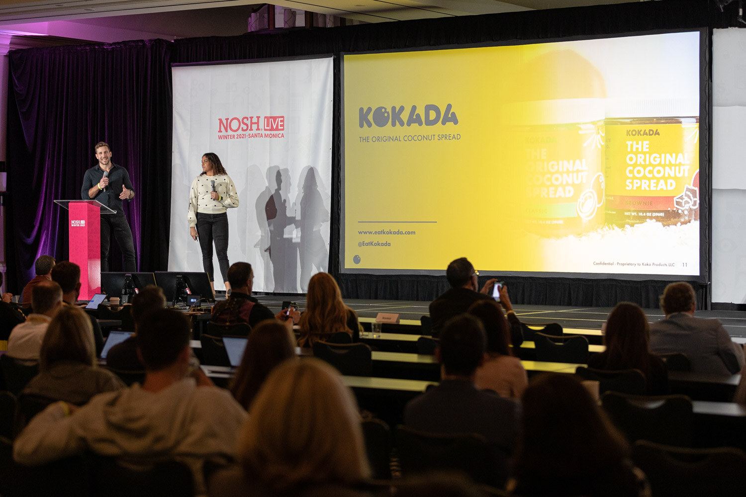 Man at a pink podium and woman in a polka-dot sweater on a stage beside a yellow screen reading Kokada The Original Coconut Spread, with audience members in the foreground.