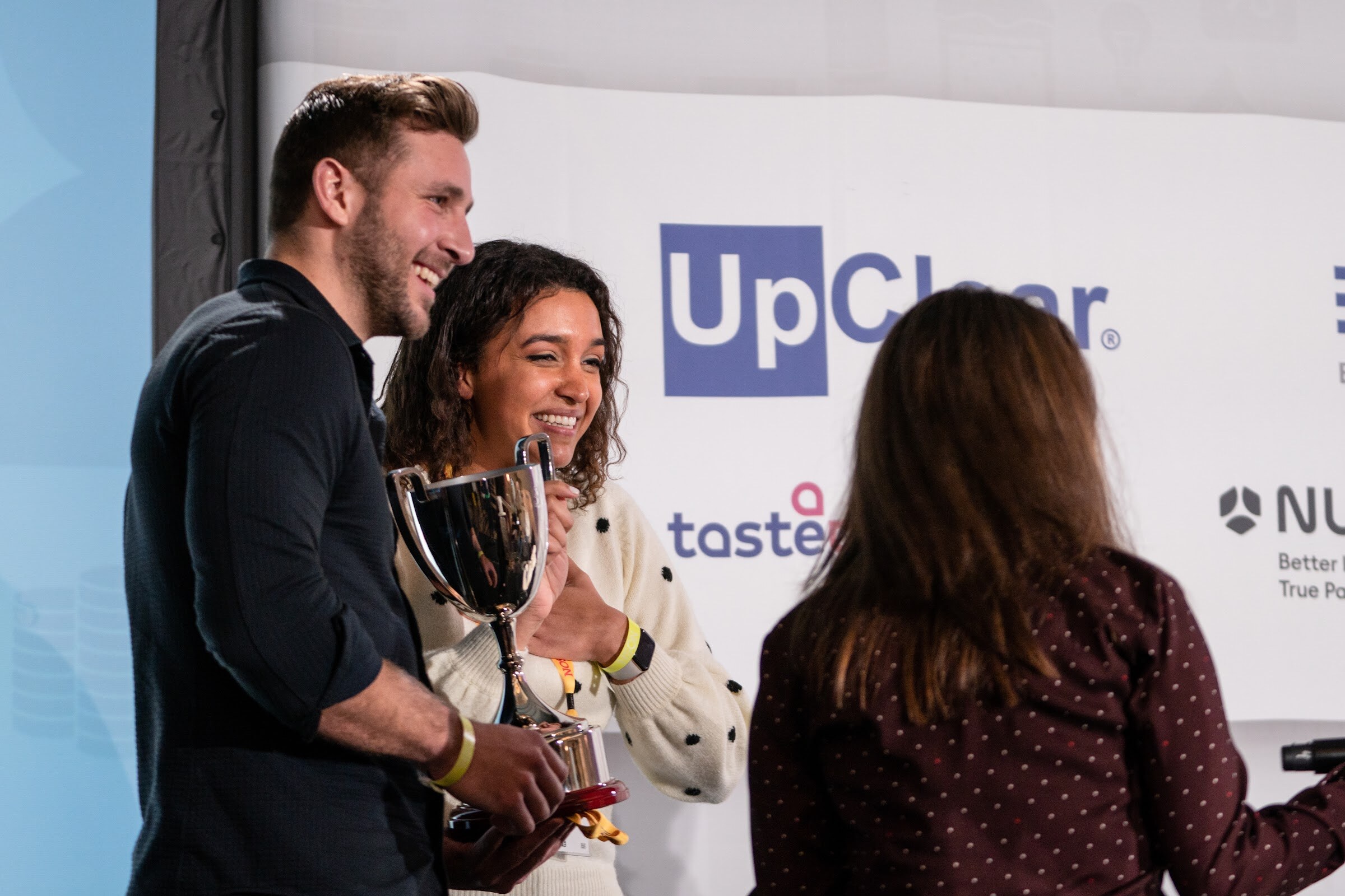 Man in a dark shirt and woman in a cream polka-dot sweater smiling while holding a silver trophy cup, with an UpClear banner behind them and a host in a polka-dot top at the right.