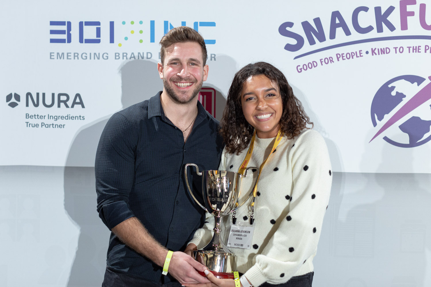 Man in a dark button-up shirt and woman in a cream polka-dot sweater smiling and holding a silver trophy cup in front of a sponsor wall with 301 Inc, NURA and SnackFutures logos.