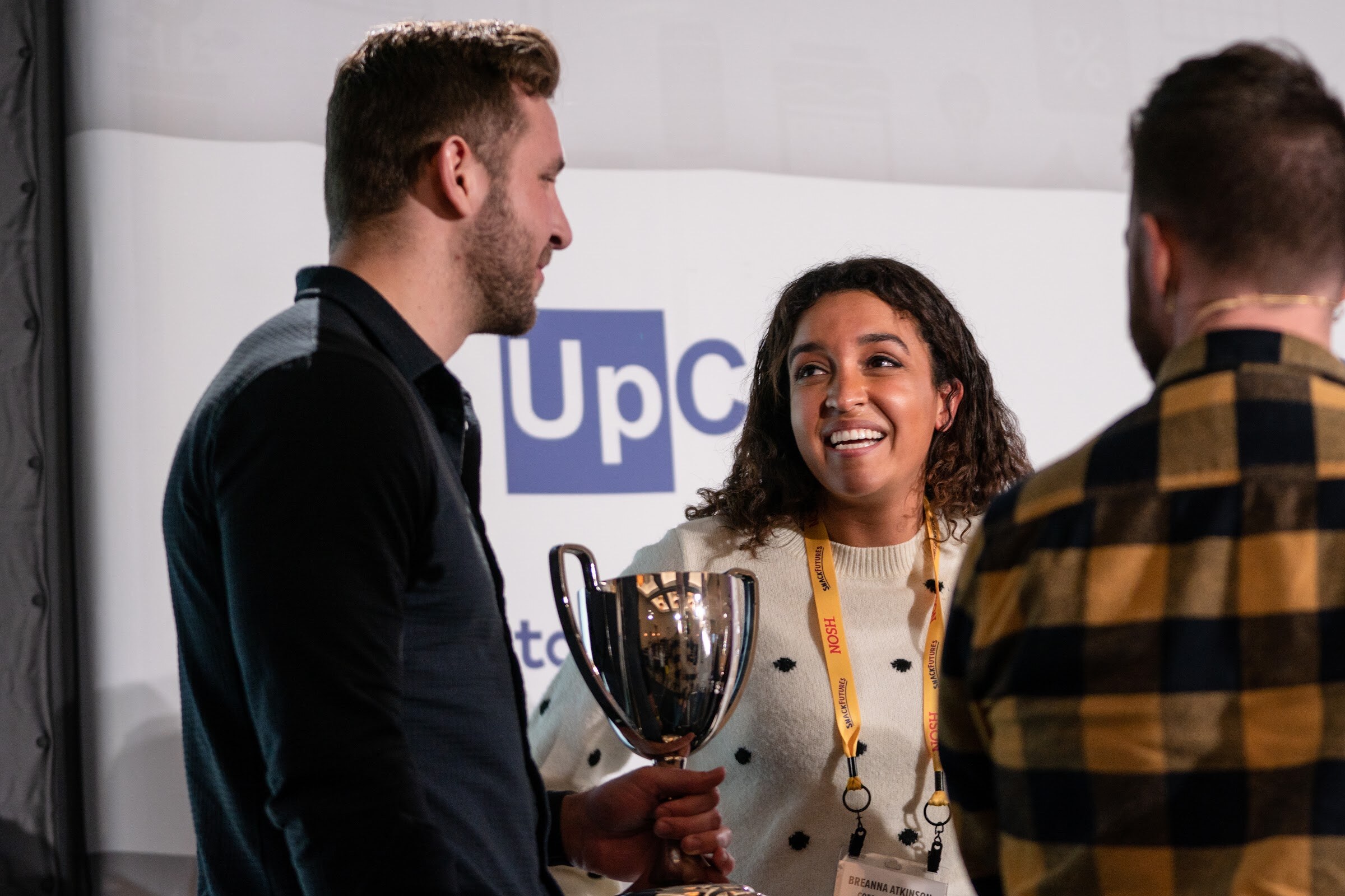 Woman with curly dark hair in a cream polka-dot sweater and a Nosh lanyard holding a silver trophy while a man in a dark shirt and a man in a plaid shirt stand beside her.