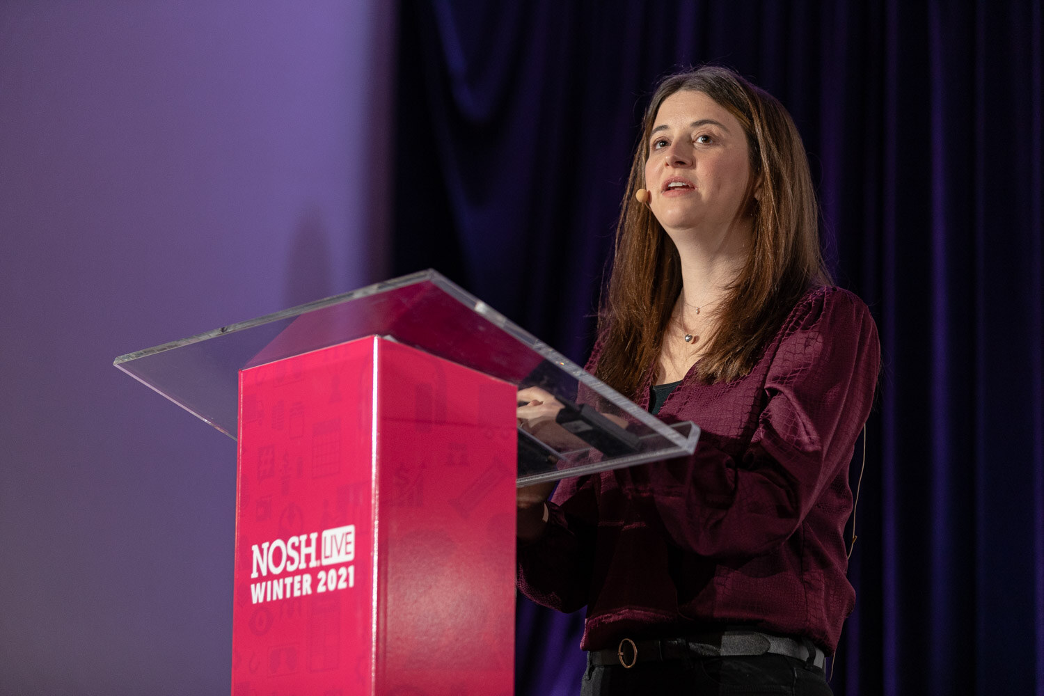 Woman with long brown hair in a burgundy blouse and headset microphone speaking at a clear podium with a pink Nosh Live Winter 2021 base.