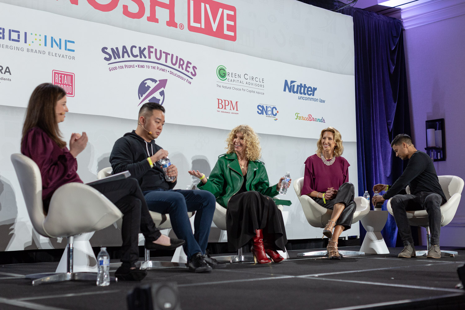 Five people seated in white chairs on stage in front of a Nosh Live sponsor wall, including a woman in a green leather jacket and red boots and a man in a black hoodie.
