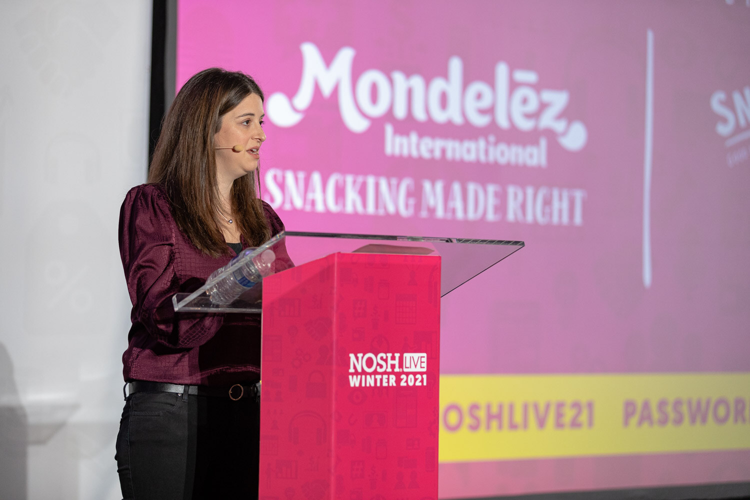 Woman in a burgundy top at a pink Nosh Live Winter 2021 podium next to a large pink screen reading Mondelez International Snacking Made Right with Wi-Fi details at the bottom.
