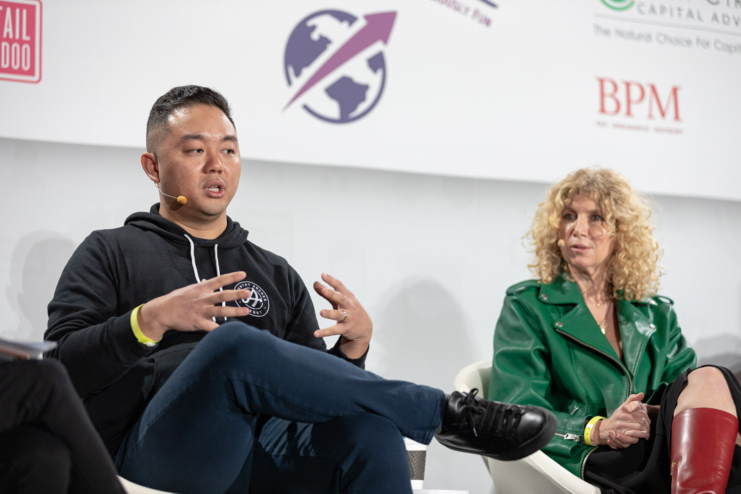 Man in a black hoodie and dark jeans gesturing with both hands beside a woman with curly blonde hair in a green leather jacket and red boots.
