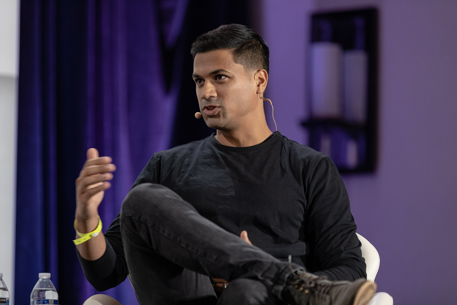 Man in a black long-sleeve shirt and boots seated in a white chair with one hand raised in front of a purple curtain.