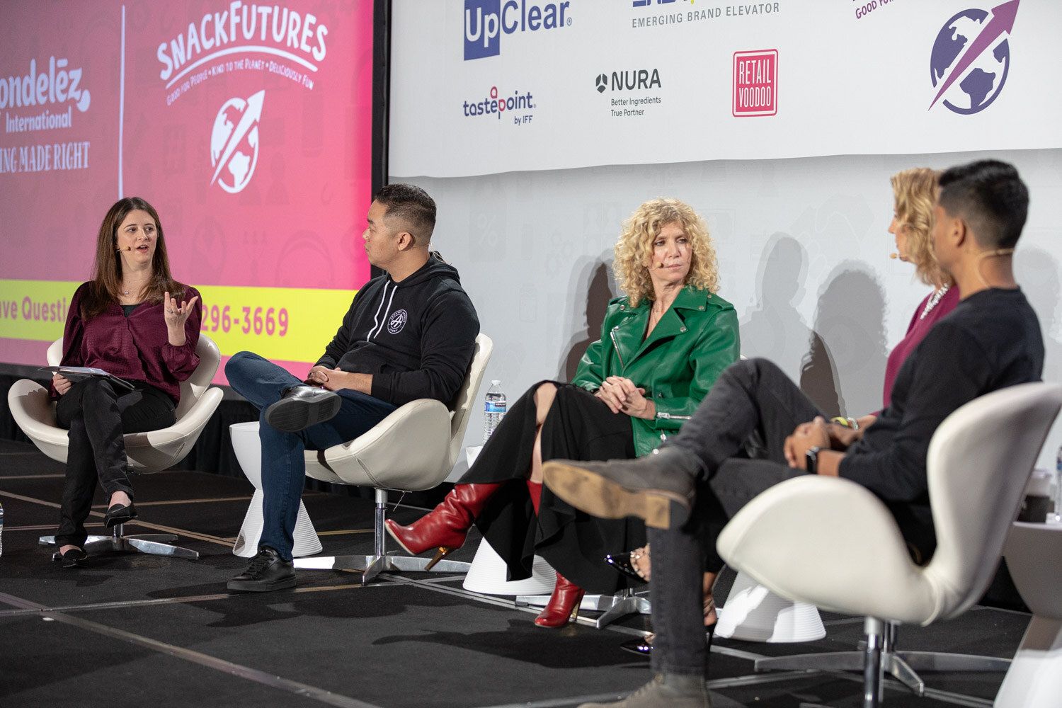 Panel of five seated in white chairs on stage next to a pink screen with Mondelez International and SnackFutures logos and a phone number for questions, in front of a sponsor wall.