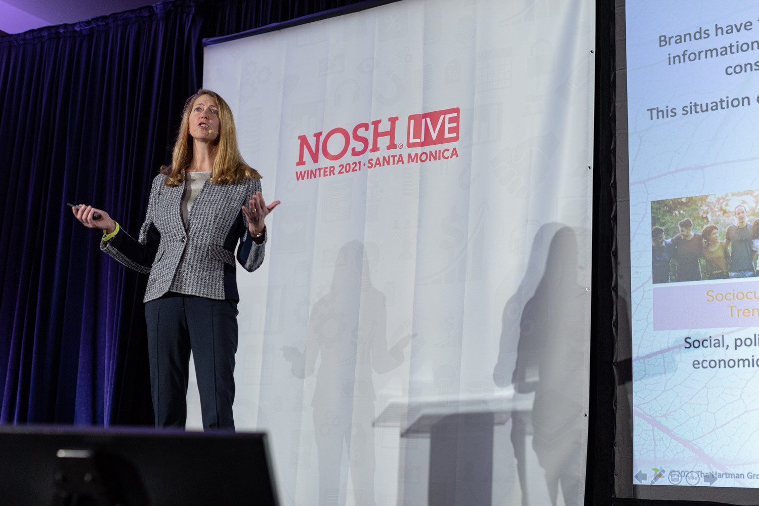 Woman in a grey and navy houndstooth blazer and navy pants standing on stage in front of a white Nosh Live Winter 2021 banner with a slide about brands and consumer information at the right.