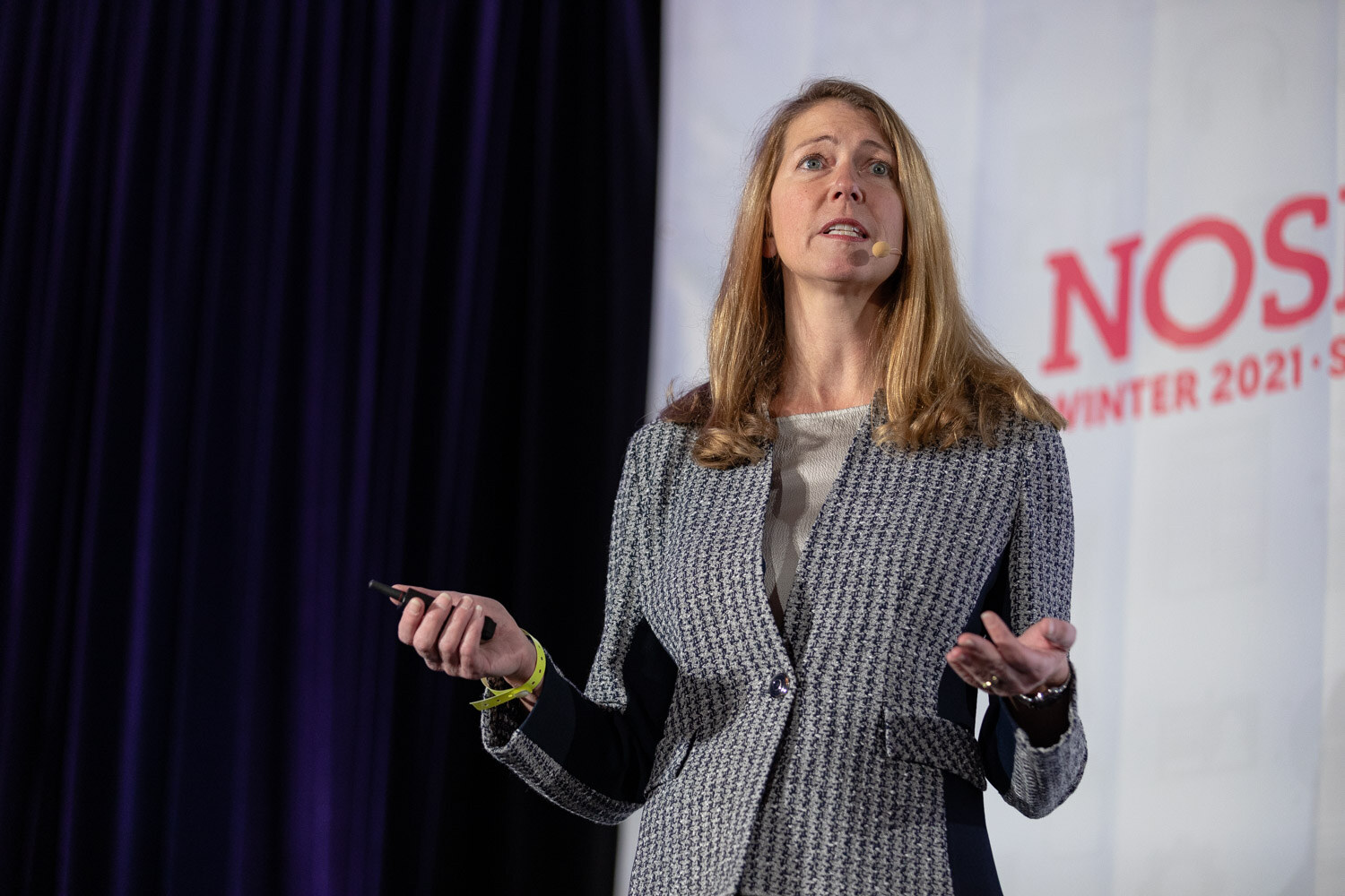Woman with shoulder-length blonde hair in a houndstooth blazer holding a slide clicker and gesturing with both hands in front of a Nosh Live banner.