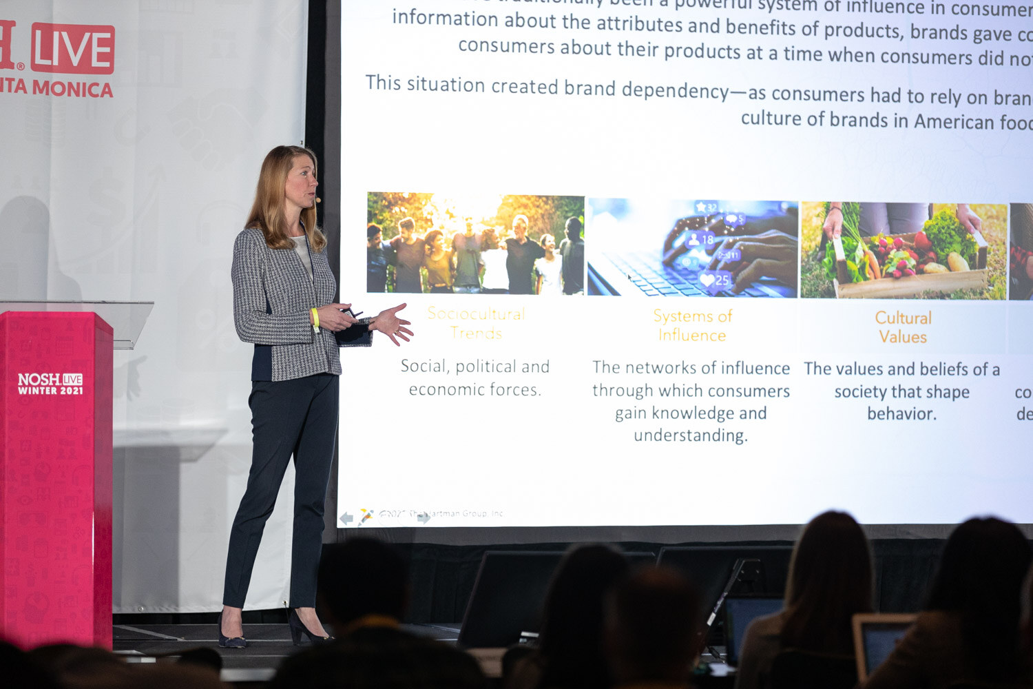 Woman in a houndstooth blazer standing next to a pink Nosh Live podium while a large slide lists Sociocultural Trends, Systems of Influence and Cultural Values.