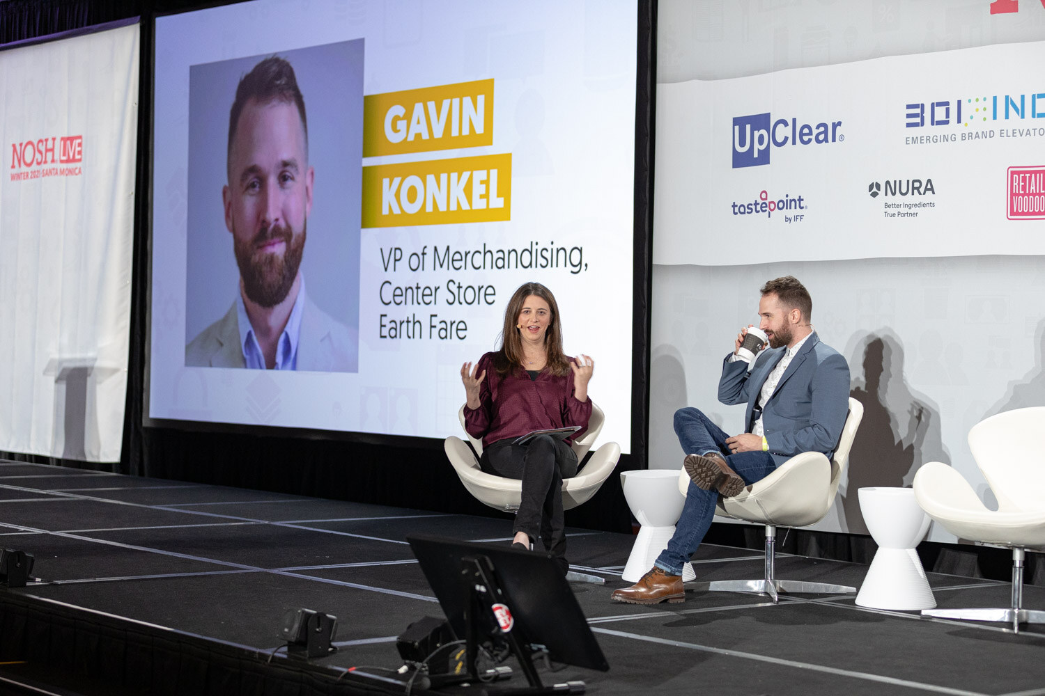 Woman in a burgundy top gesturing and a bearded man in a blue blazer sipping coffee on white chairs, with a large slide showing his portrait and the text VP of Merchandising, Center Store, Earth Fare.