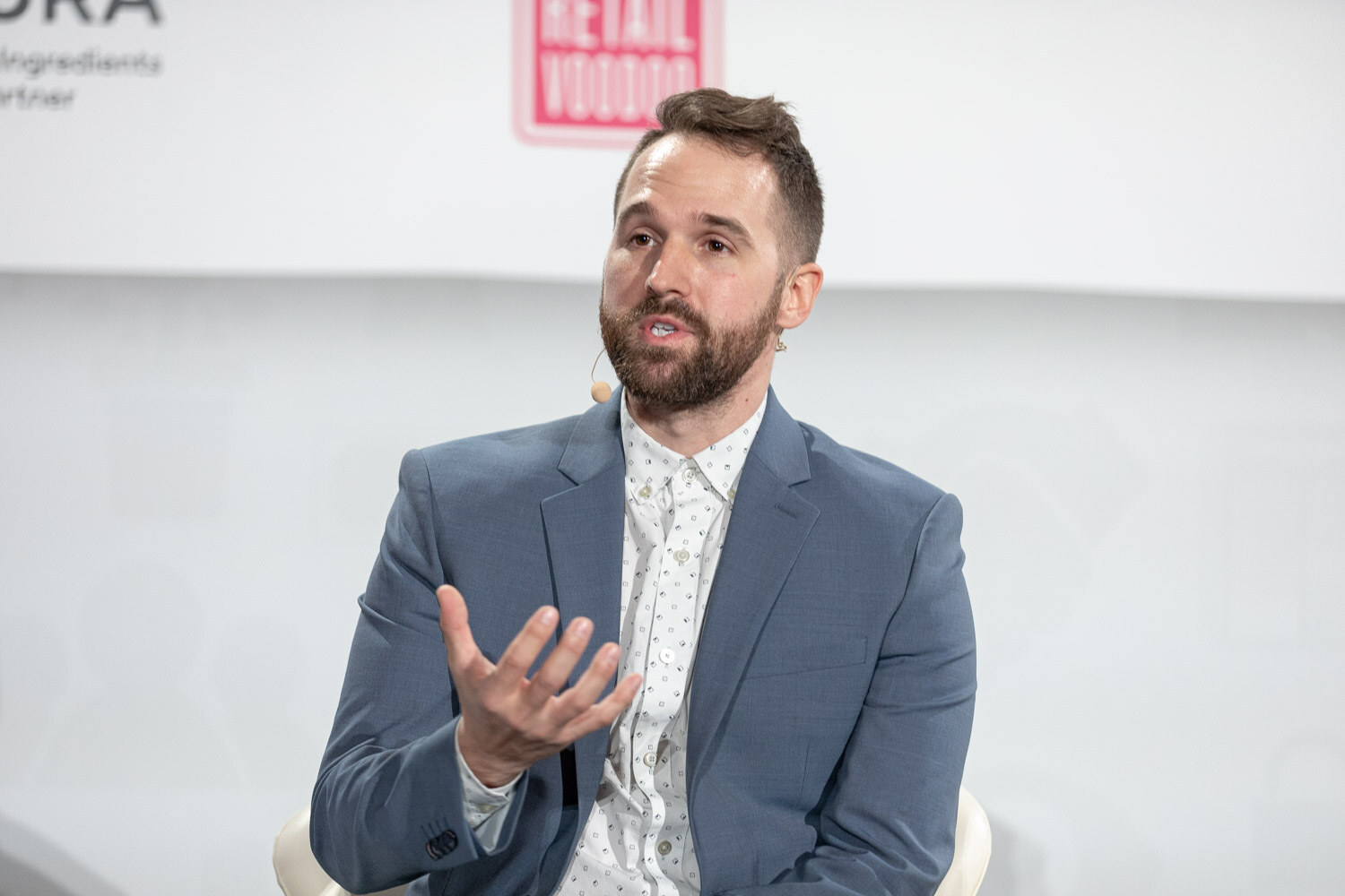Bearded man in a light blue blazer and white patterned shirt gesturing with one hand, seated in front of a white backdrop with NURA and Retail Voodoo logos.