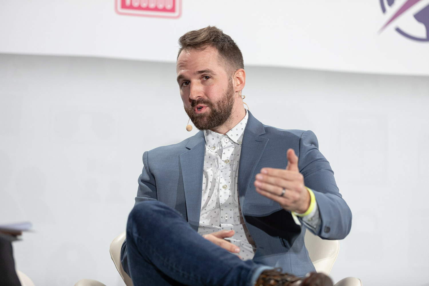 Bearded man in a light blue blazer and jeans, with a yellow wristband, gesturing with an open hand in a white chair in front of a Retail Voodoo logo.