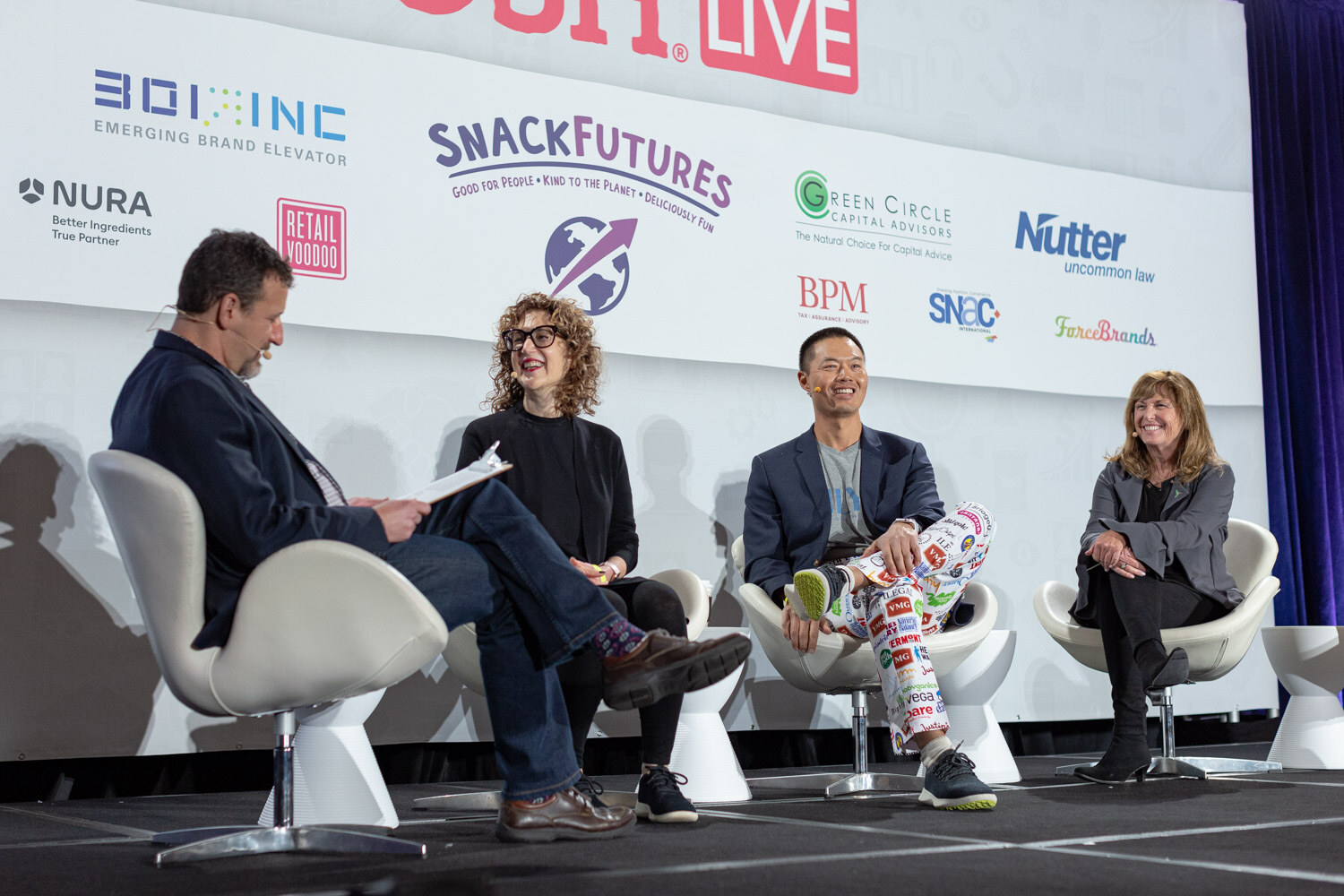Four people seated in white chairs on a stage in front of sponsor logos, including a man in a navy blazer and brightly printed pants covered with brand logos.