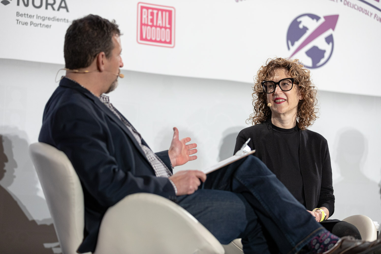 Man in a navy blazer holding a clipboard turned toward a woman with curly hair and bold black glasses in a black top, seated on stage in front of NURA and Retail Voodoo logos.