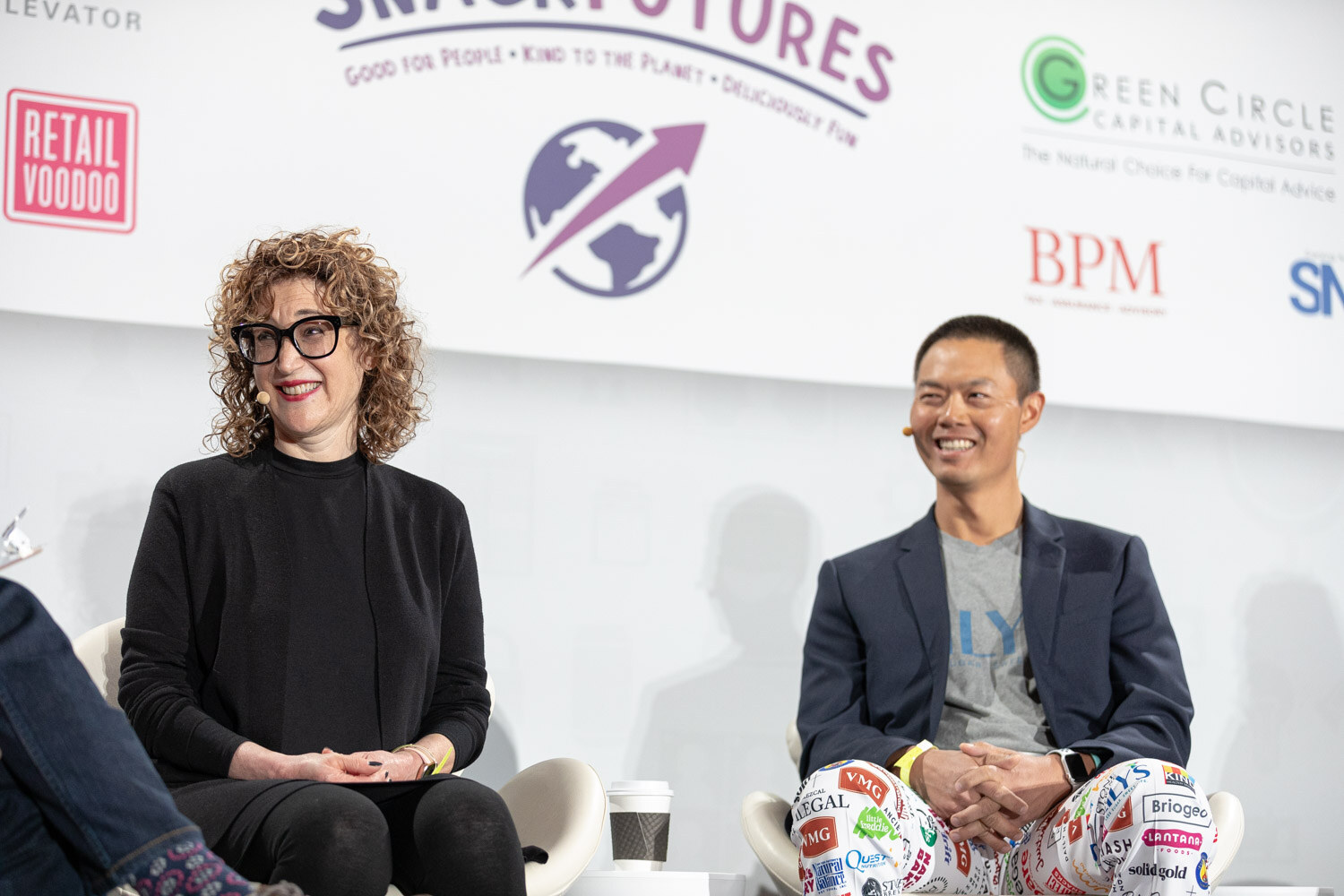 Woman with curly hair and black glasses in a black sweater and a smiling man in a navy blazer and brand-logo patterned pants seated on white chairs.