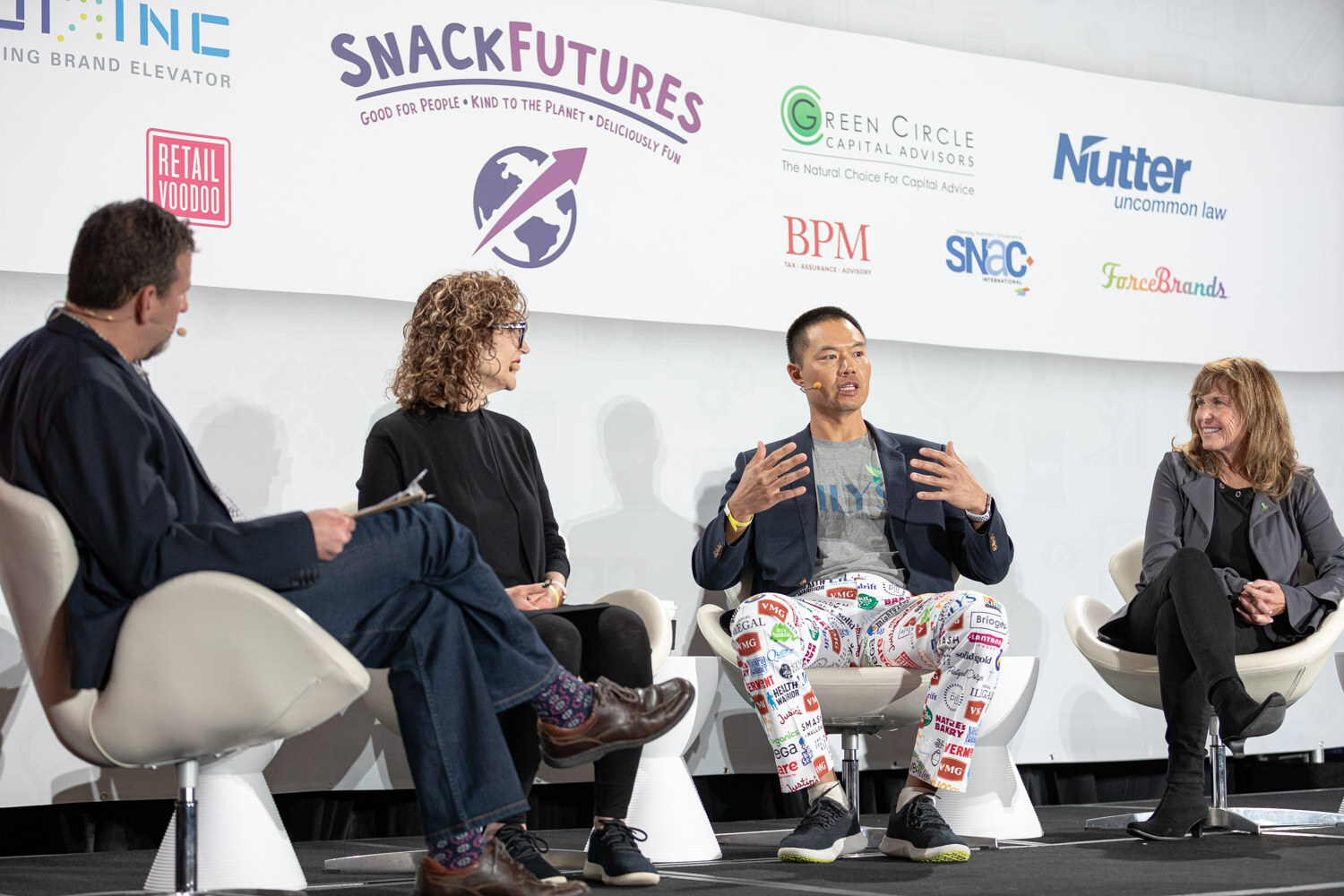 Four people on stage in white chairs with a man in a navy blazer and logo-patterned pants gesturing in front of a SnackFutures, Green Circle Capital Advisors and Nutter sponsor wall.