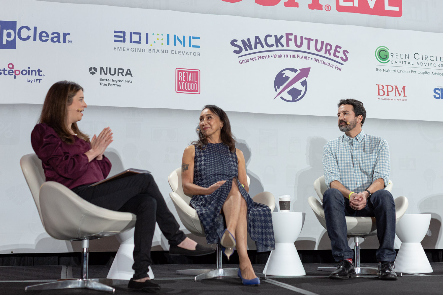 Woman in a burgundy top applauding while seated on a white chair beside a woman in a houndstooth dress and a man in a gingham shirt, in front of a sponsor wall with Tastepoint, NURA and Retail Voodoo logos.