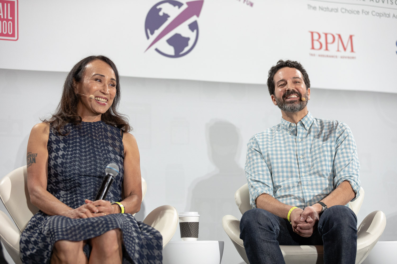 Woman in a navy houndstooth dress holding a microphone and a bearded man in a blue gingham shirt both smiling widely on white chairs.