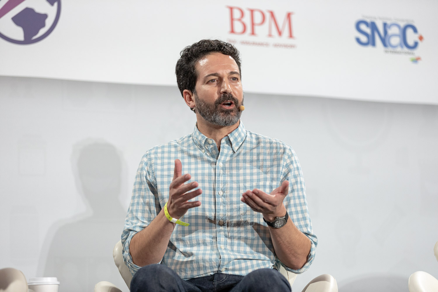 Man with dark curly hair and a grey beard in a blue gingham shirt gesturing with both hands in front of BPM and SNAC logos.