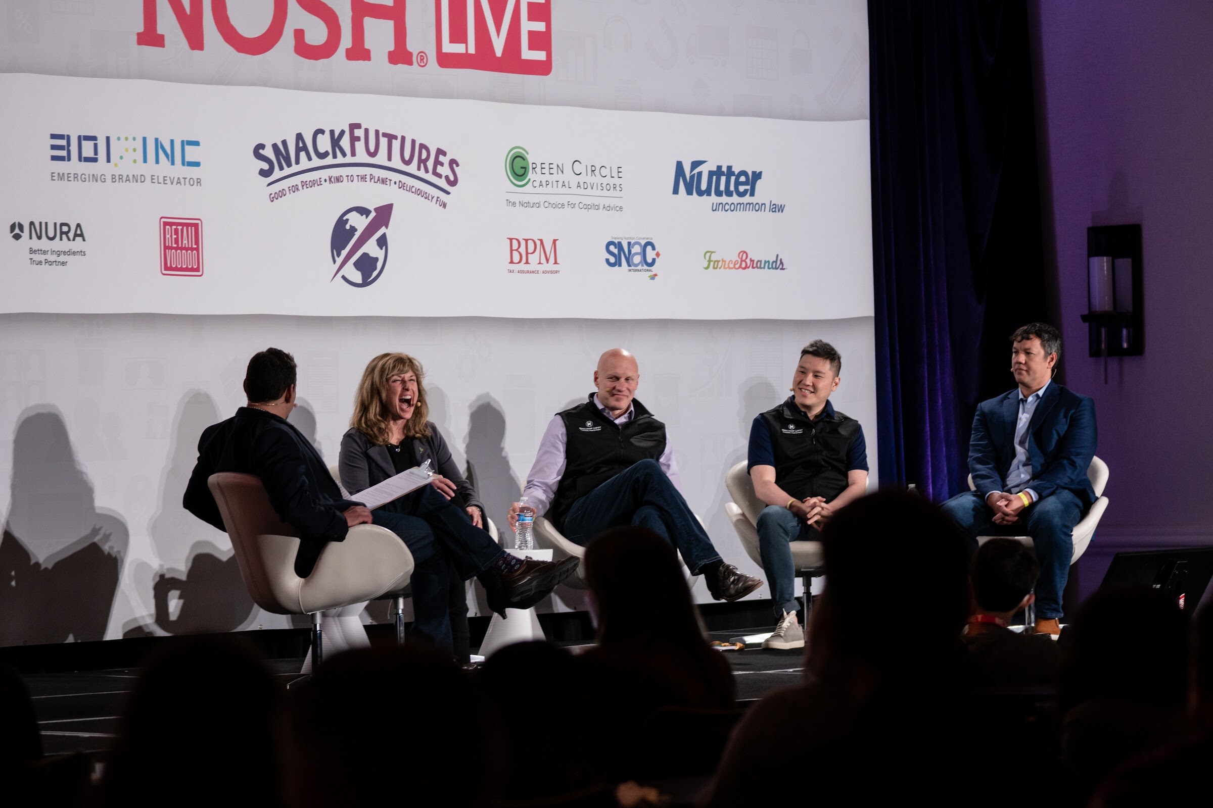 Five people seated on a stage in front of the Nosh Live sponsor wall, including a woman laughing, a bald man in a black vest and a man in a navy blazer.