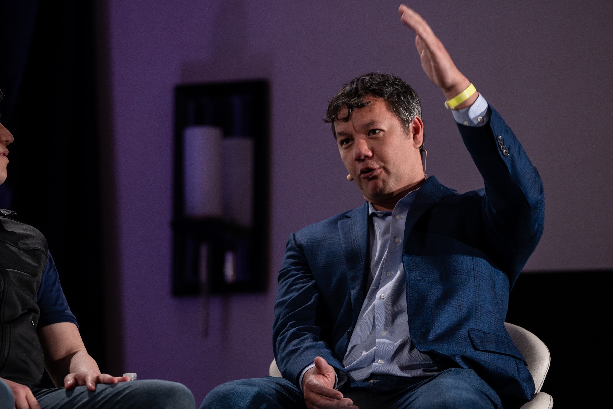 Man in a navy plaid blazer and light blue shirt with a yellow wristband raising one hand in front of a dark purple curtain.