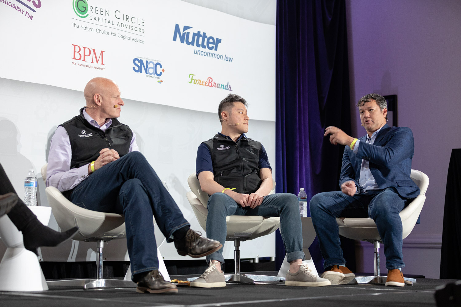 Three men on white chairs on stage, two in black vests and one in a navy blazer gesturing, in front of Green Circle Capital Advisors, Nutter, BPM, SNAC and ForceBrands logos.