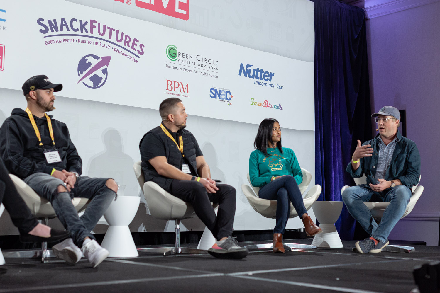 Four people seated in white chairs on stage, two men in dark jackets with Nosh lanyards, a woman in a teal sweatshirt and a man in a blue cap, in front of SnackFutures and Nutter logos.