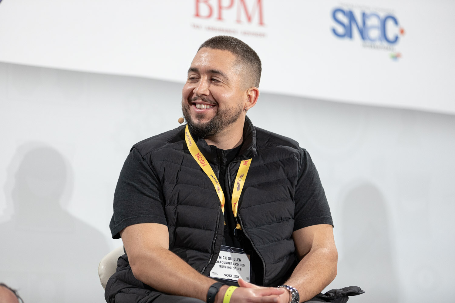 Smiling bearded man in a black puffer vest and yellow Nosh lanyard, with a badge that reads Truff Hot Sauce, in front of BPM and SNAC logos.