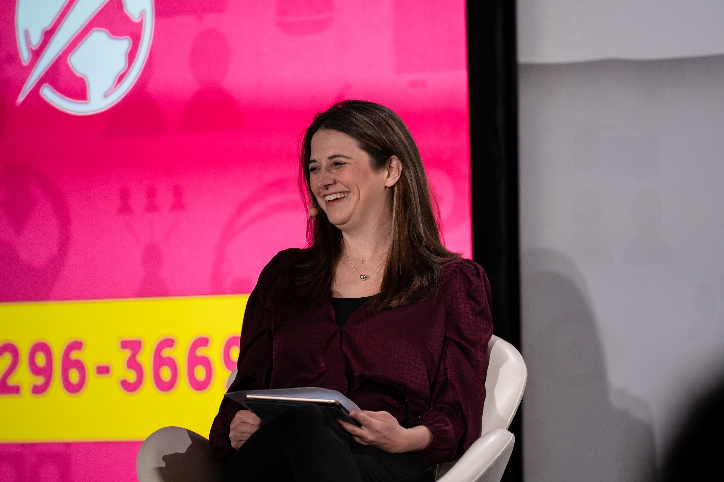 Woman in a burgundy top holding a tablet and laughing, seated in a white chair in front of a pink screen with part of a phone number.