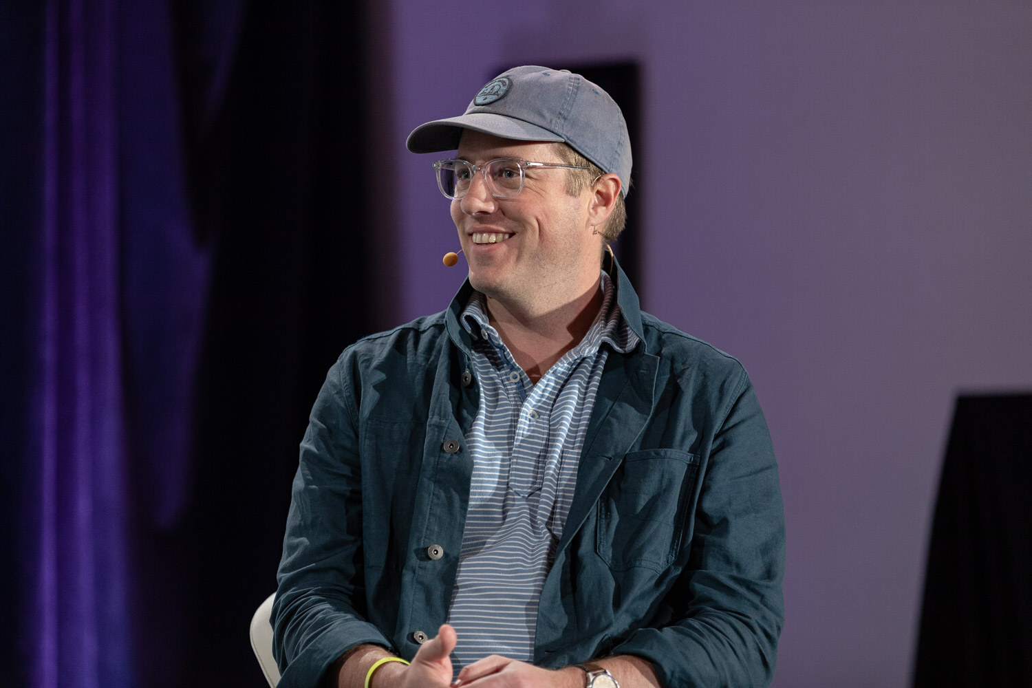 Man in a grey baseball cap, clear-framed glasses, a dark green jacket and a striped polo smiling on a stage with a purple curtain behind him.