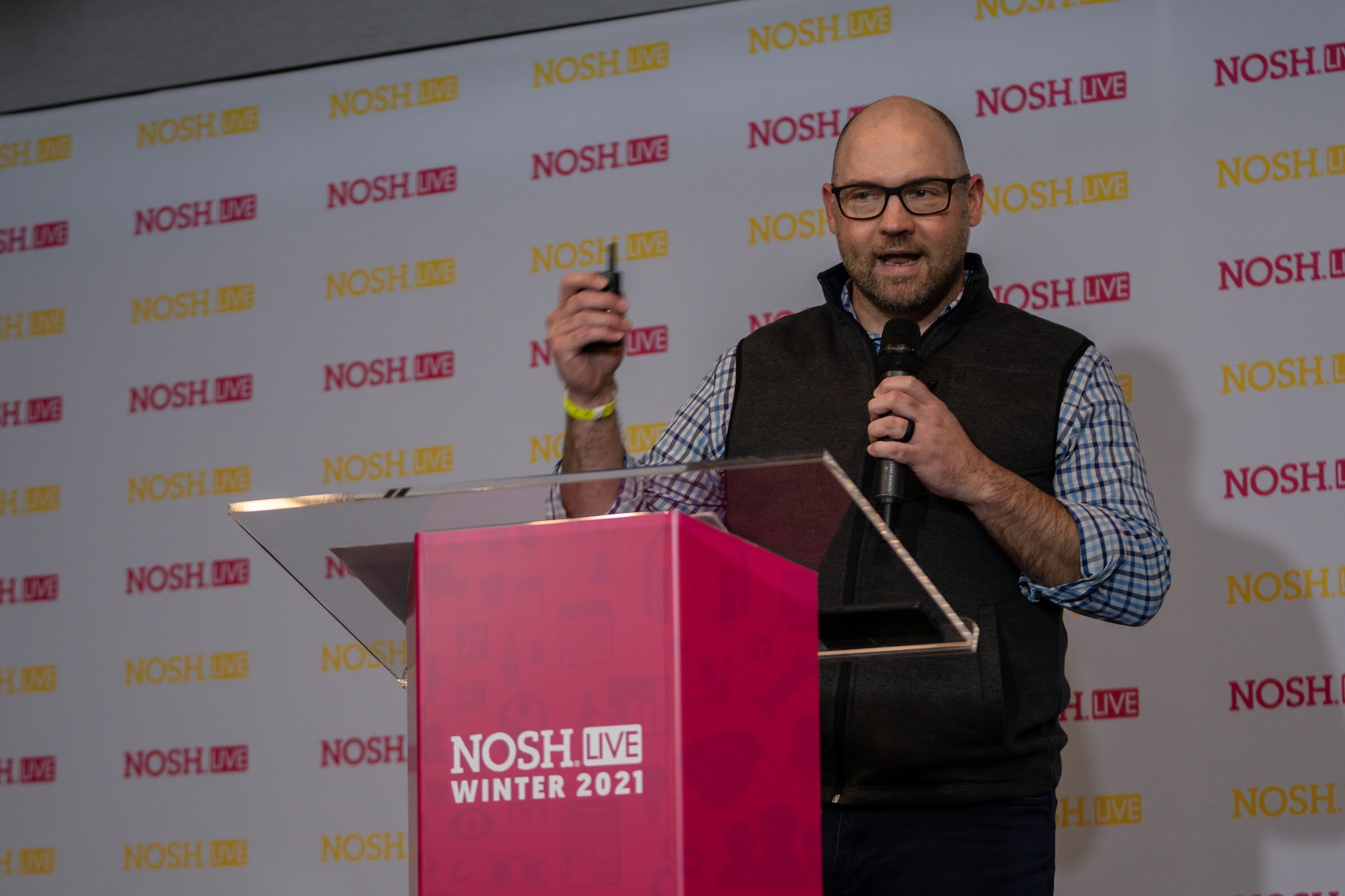 Bald man with glasses in a dark fleece vest and checked shirt holding a clicker and microphone behind a pink Nosh Live Winter 2021 podium in front of a repeating Nosh Live backdrop.