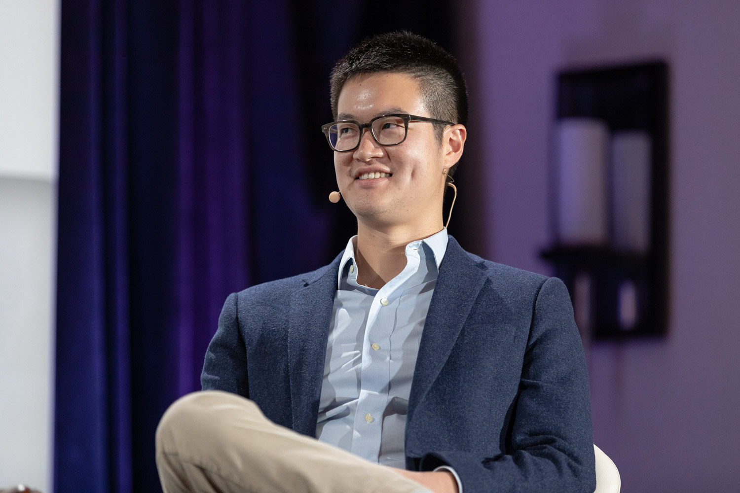 Man with dark-framed glasses and a headset microphone in a navy blazer over a light blue shirt, seated in front of a dark curtain.