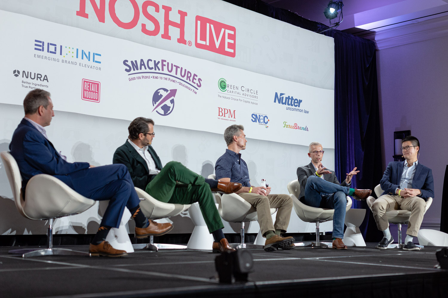 Five men seated in white chairs on stage, in navy suit, green pants and blazer, in front of the Nosh Live sponsor wall.