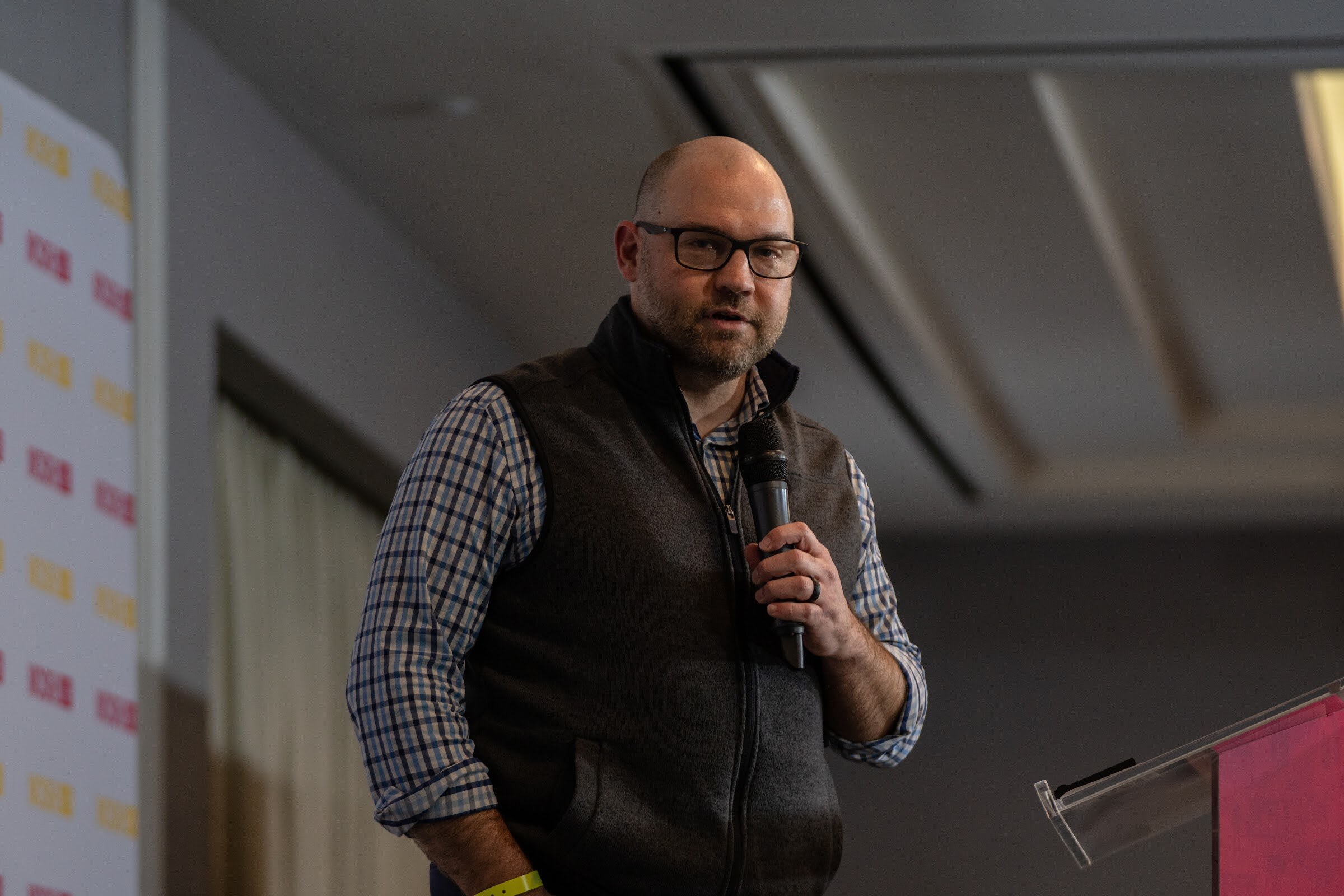 Bald man with glasses and a beard in a dark vest over a checked shirt holding a microphone, with a clear podium and a Nosh Live backdrop at the left.