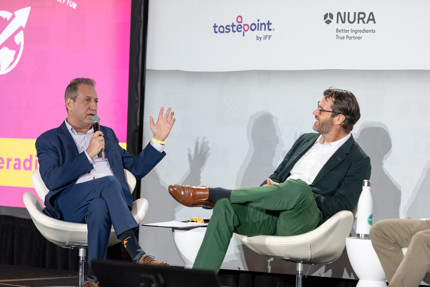 Man in a navy blazer holding a microphone and gesturing while a bearded man in green pants and a dark jacket turns toward him, with a pink screen and Tastepoint and NURA logos behind.