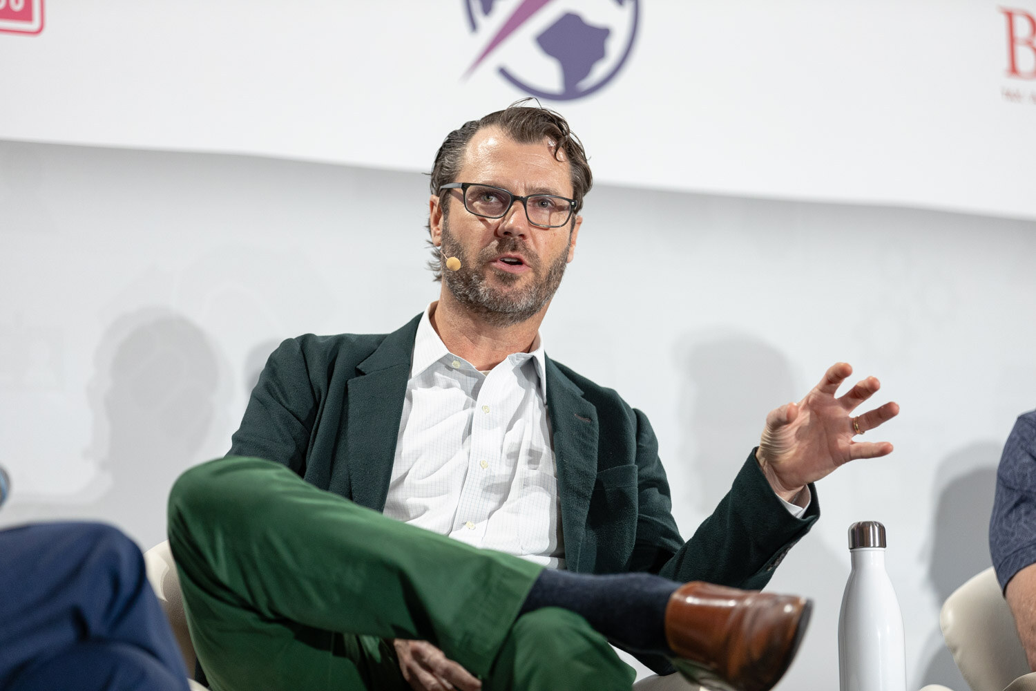 Man with glasses and a beard in a dark green blazer and green pants, seated and gesturing, with a white water bottle beside him.