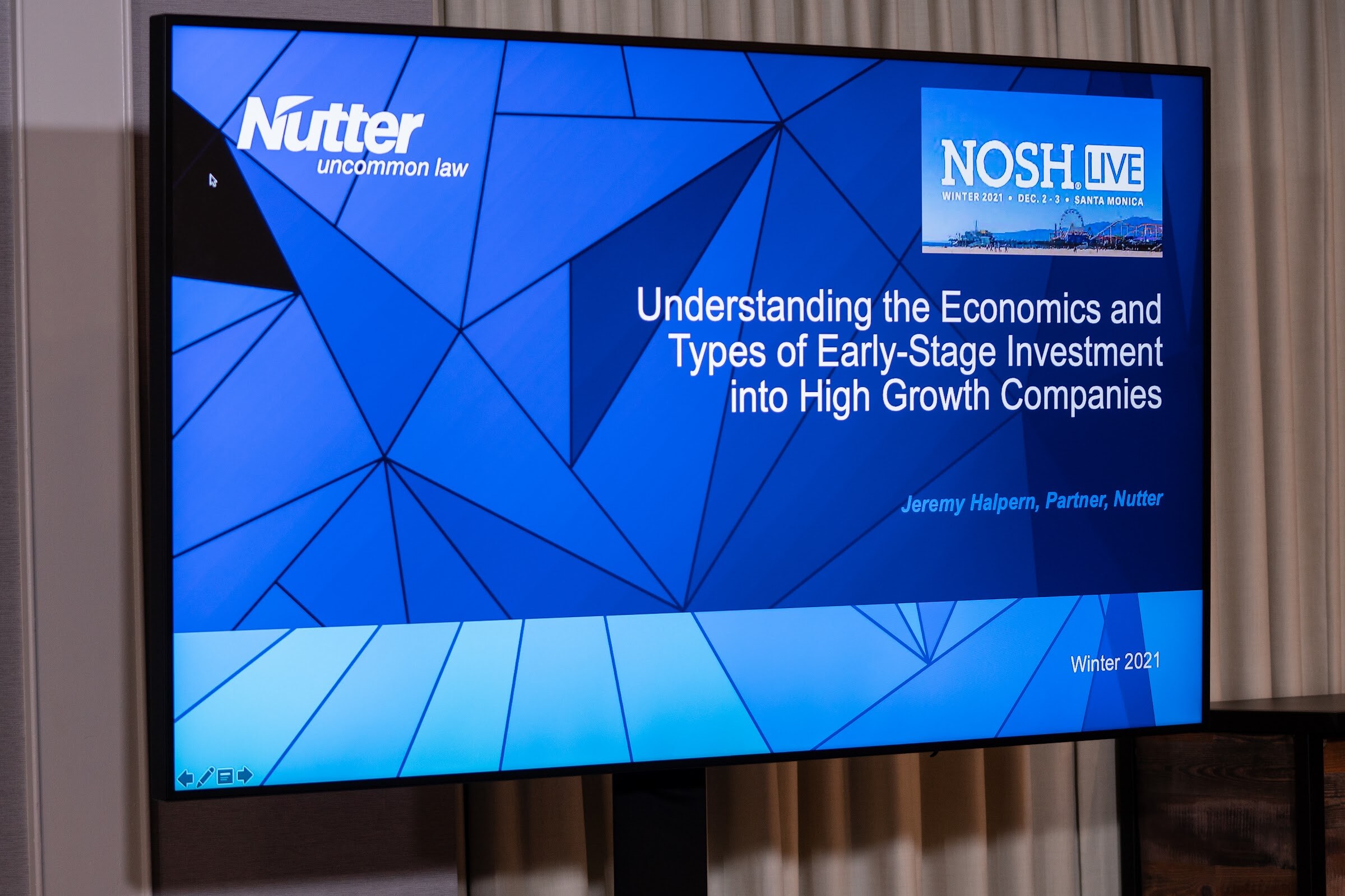 Wall-mounted screen showing a blue Nutter slide titled Understanding the Economics and Types of Early-Stage Investment into High Growth Companies with the Nosh Live Winter 2021 Dec 2-3 Santa Monica logo.
