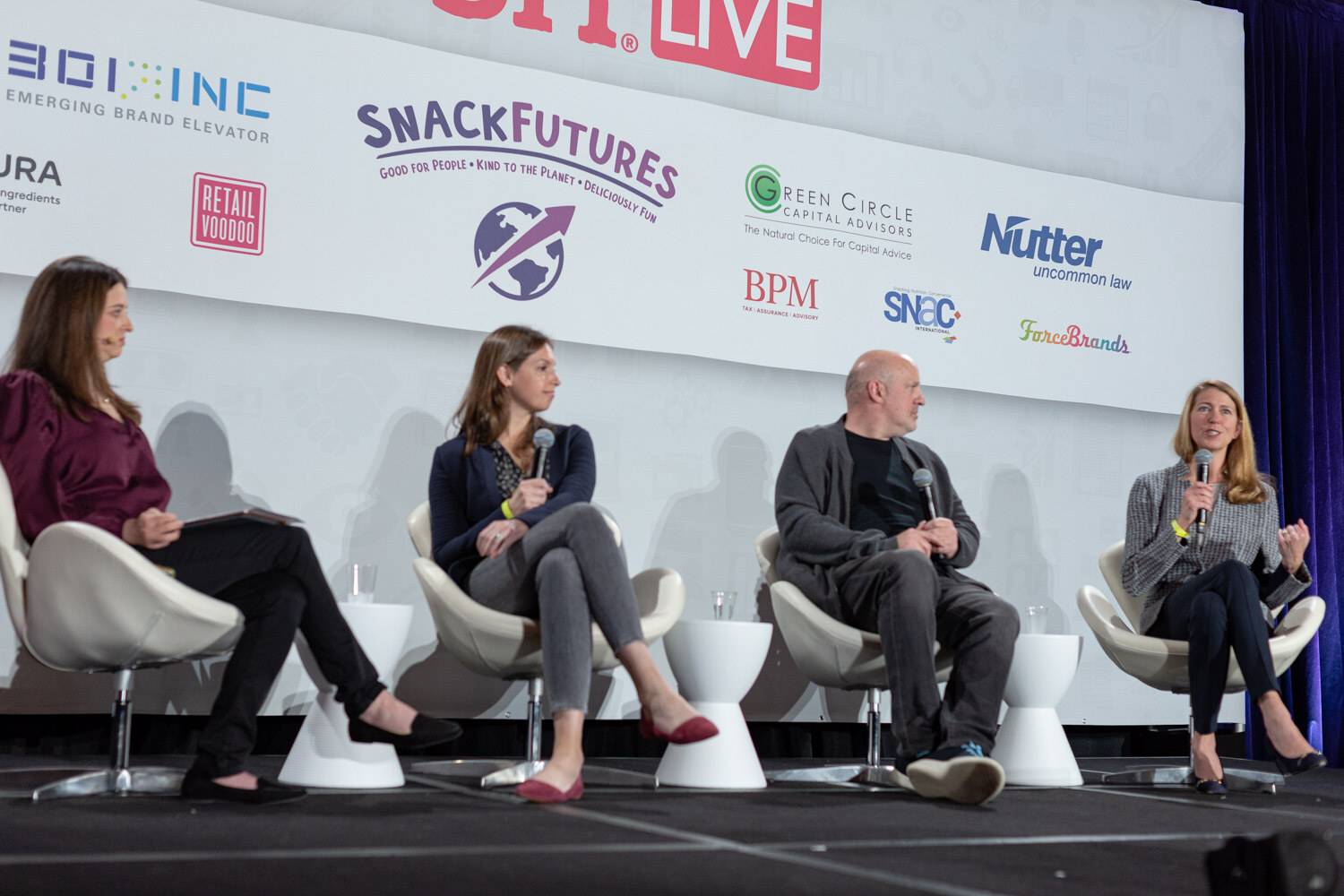 Four women and men seated in white chairs on stage in front of the Nosh Live sponsor wall, with the moderator in a burgundy top at the left and a woman in a houndstooth blazer speaking at the right.