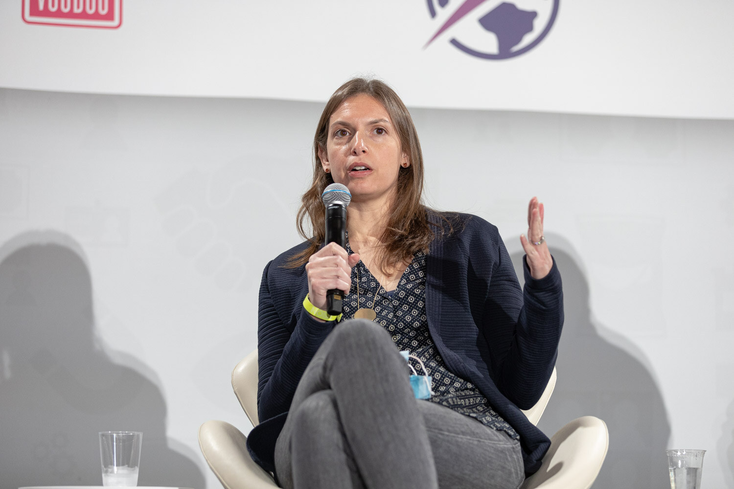 Woman with long brown hair in a navy cardigan over a patterned blouse holding a microphone and gesturing, seated in a white chair with a face mask hanging from her neck.